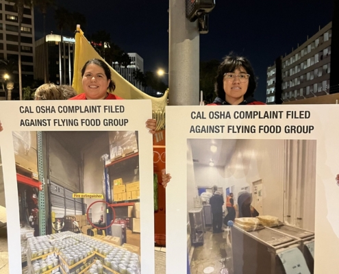 Two women stand under a street light in the evening holding up photo enlargements from a safety complaint