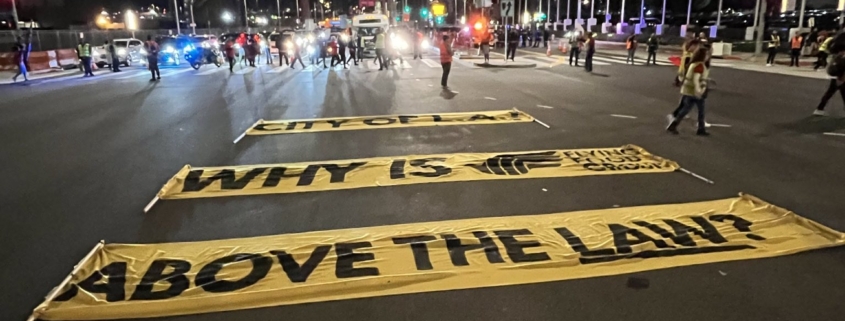 Large yellow banners lie in the center of the busy intersection at Century Boulevard and Avion Drive near LAX. The banners read, "City of LA: Why is Flying Food Group above the law?"