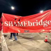 Two hotel workers stand 10 feet apart to display a large red vinyl banner with the SHAMEBridge logo