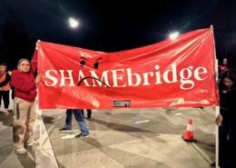 Two hotel workers stand 10 feet apart to display a large red vinyl banner with the SHAMEBridge logo