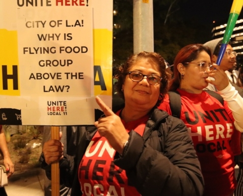 A woman stands outside under a streetlight in the evening pointing to her sign that reads "City of LA: Why is Flying Food Group above the law?"