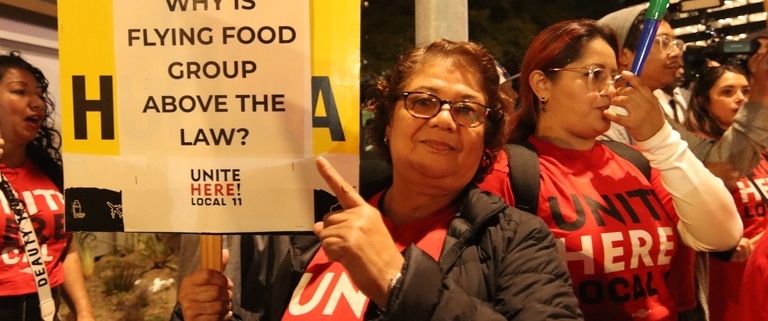 A woman stands outside under a streetlight in the evening pointing to her sign that reads "City of LA: Why is Flying Food Group above the law?"