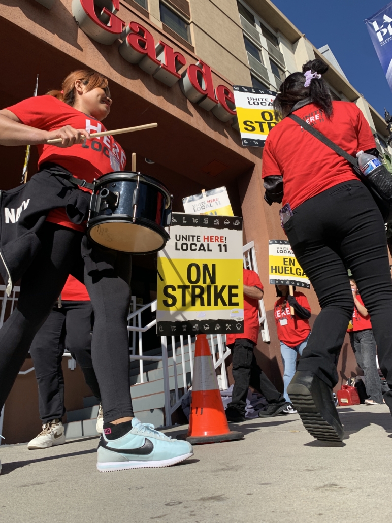 Looking up from the sidewalk outside a hotel to see traffic cones, signs that read "UNITE HERE Local 11 ON STRIKE," and two women in red T-shirts with drums and drumsticks