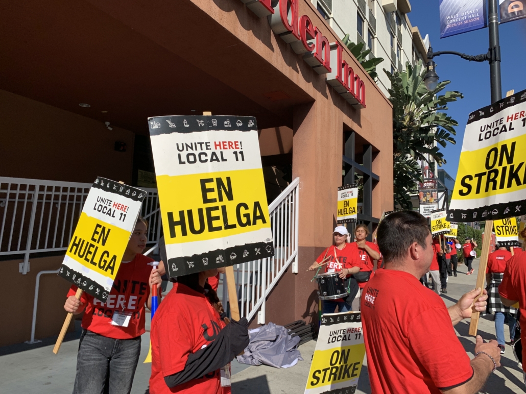 Outside a brown hotel building, workers with red t-shirts and drums picket with signs reading "UNITE HERE Local 11 ON STRIKE" and "UNITE HERE Local 11 EN HUELGA"