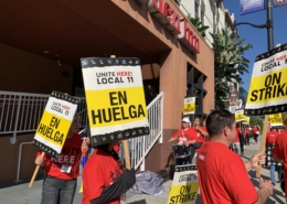 Outside a brown hotel building, workers with red t-shirts and drums picket with signs reading "UNITE HERE Local 11 ON STRIKE" and "UNITE HERE Local 11 EN HUELGA"