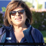 A woman with shoulder-length brown hair in sunglasses and a blue work uniform top speaks into the camera