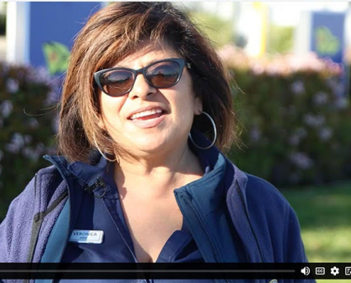 A woman with shoulder-length brown hair in sunglasses and a blue work uniform top speaks into the camera