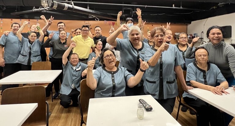 A group of two dozen hotel housekeepers in blue uniforms smile and raise their arms in victory as they learn about their new union contracts
