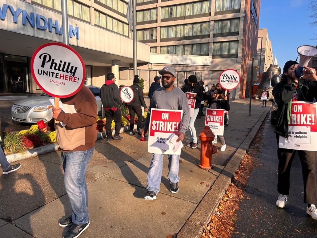 On a beautiful November day outside a building marked "Wyndham" in blue letters, striking workers picket with signs reading "ON STRIKE! at Wyndham Philadelphia" and "Philly UNITE HERE Local 274"