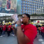 In the foreground, an African American man chants into a bullhorn. In the background, hotel workers picket outside a Santa Monica hotel