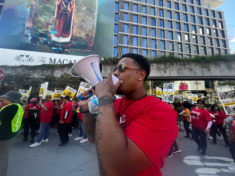 In the foreground, an African American man chants into a bullhorn. In the background, hotel workers picket outside a Santa Monica hotel