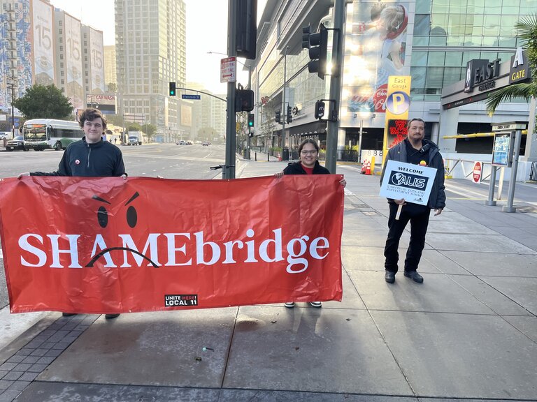 Two activists stand on a downtown Los Angeles sidewalk holding a 10-foot wide red banner with the coinage "SHAMEBridge" printed on it