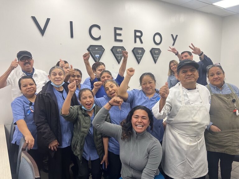 A group of a dozen hotel workers smile and raise their arms in victory in front of a wall that reads "Viceroy"