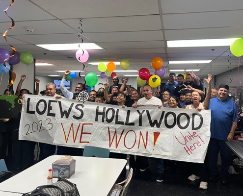 A group of 50 hotel workers with balloons stand behind a banner that reads "Loews Hollywood: We Won!"